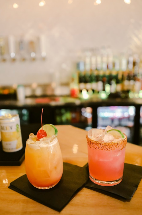 Two colorful cocktails on a bar counter, one garnished with a cherry and lime, the other with a salted rim.